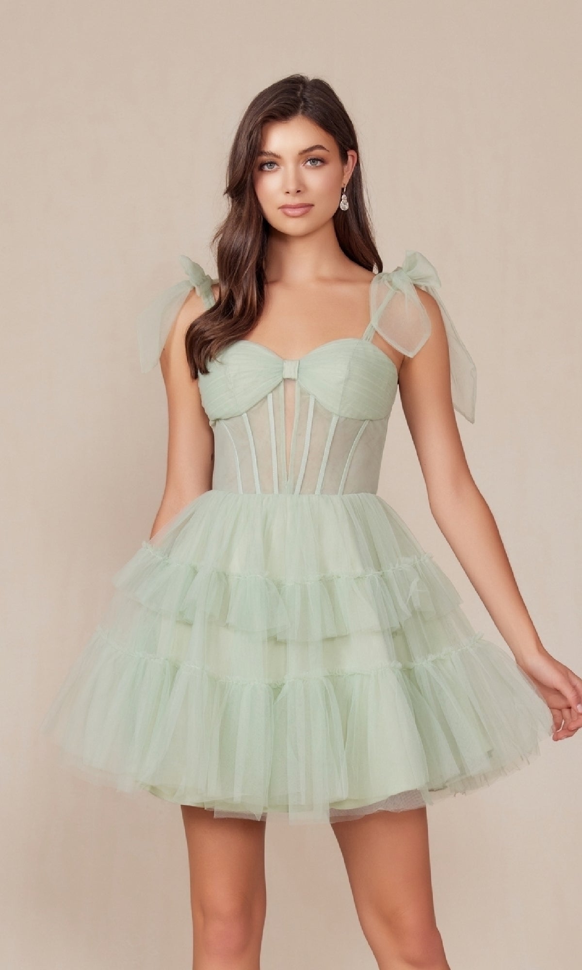 Woman wearing the Shoulder-Tie Short Ruffled A-Line Hoco Dress Y858, featuring a light green tiered tulle skirt, sheer corset bodice, and ribbon shoulder straps, posing against a plain beige background.